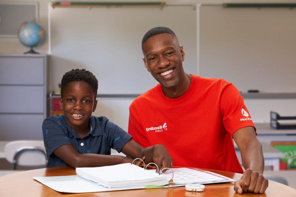 Seated side-by-side, an adult in a red shirt and a child in a blue shirt smile at the camera. They lean on a table with an open book.